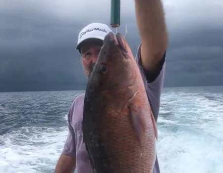 fisherman holds up his catch with one hand on a boat with a cloudy sky.